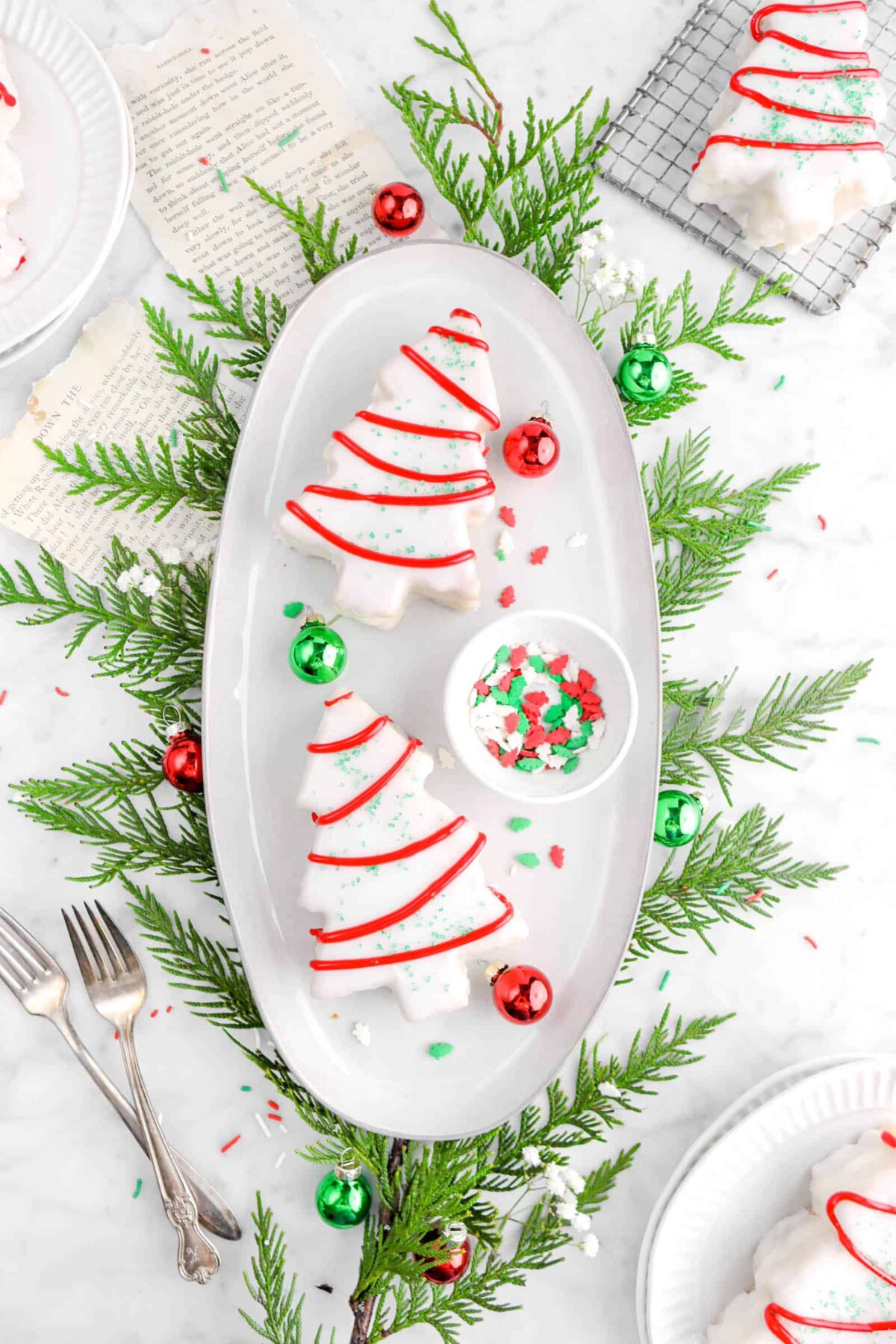 two christmas tree cakes on white plate with greenery underneath, two forks, three christmas cakes around, two book pages underneath plate, and mini red and green ornments around