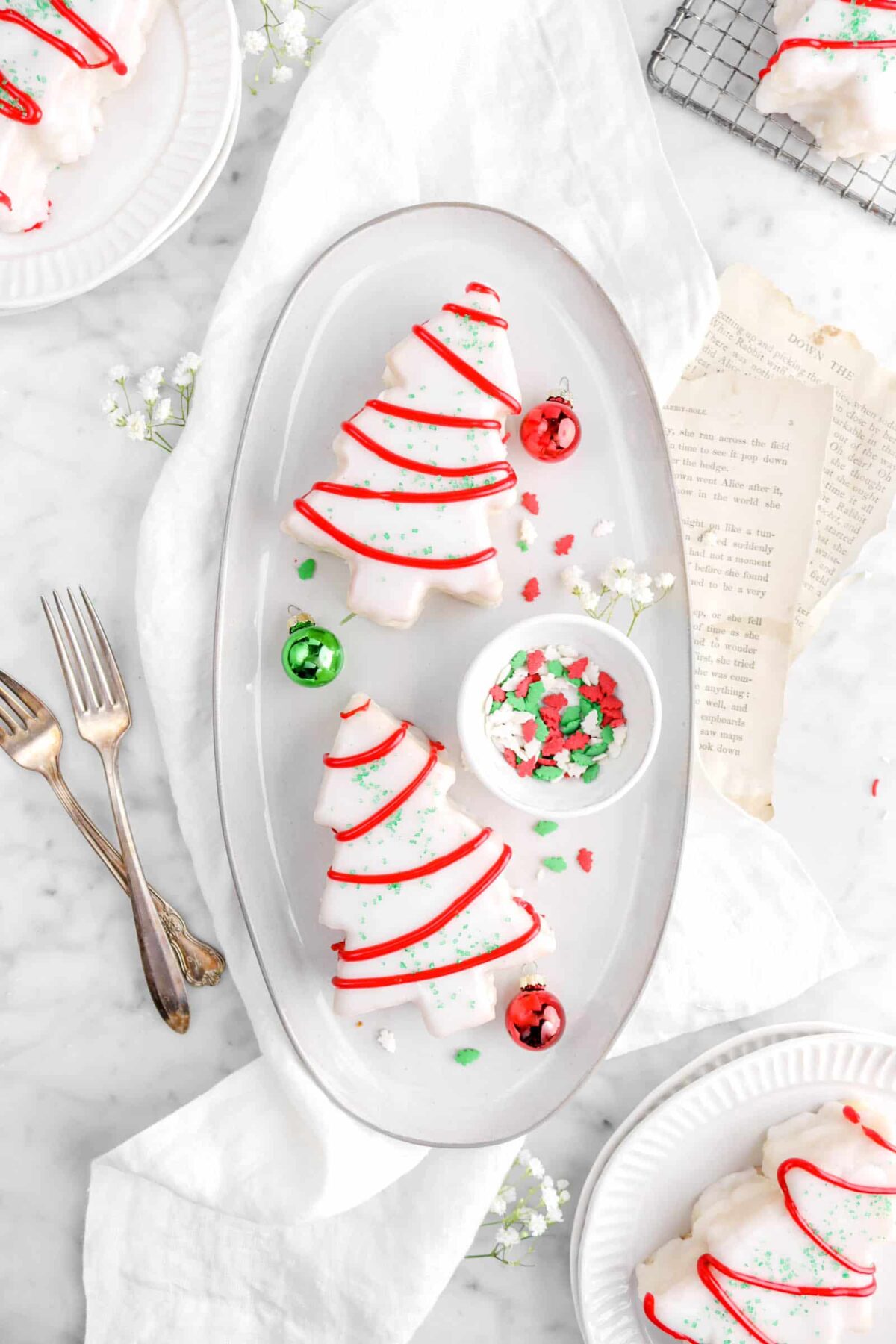two cakes on serving plate with sprinkles, red and green ornaments, and flowers on top of a white napkin with two forks, book pages, and plates