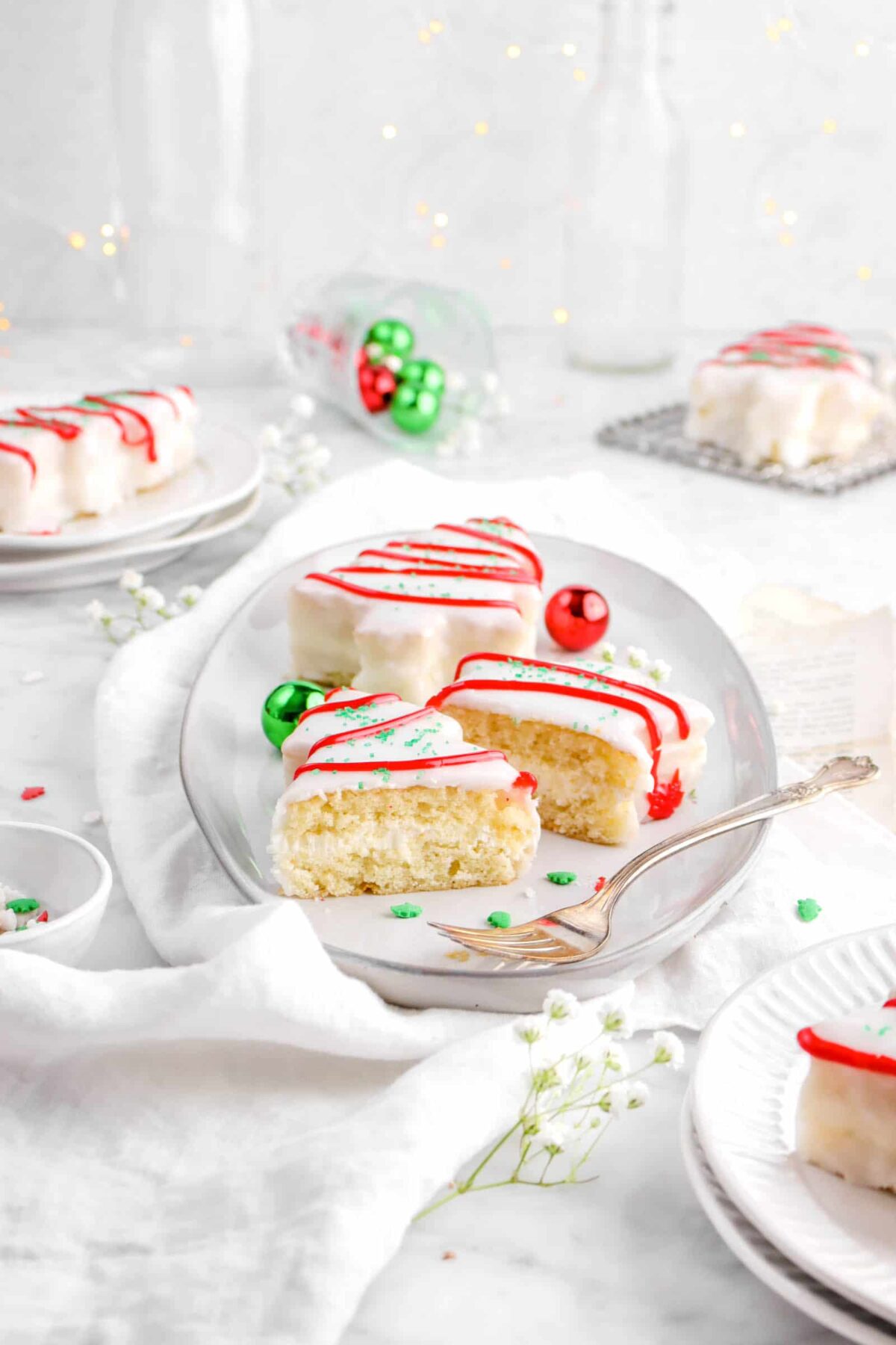 pulled back photo of christmas tree cakes on white plate with one cut in half with fork in front, fairy lights behind and ornaments