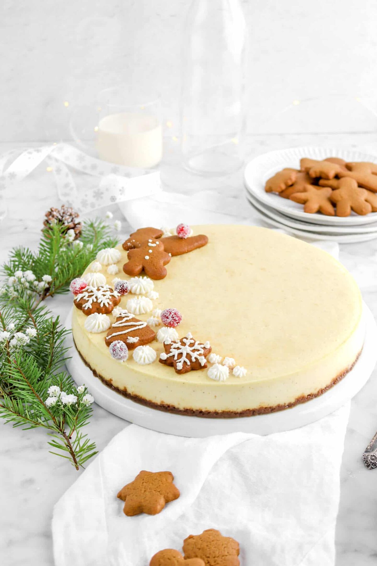eggnog cheesecake on plate on top of white napkin with plate of cookies behind, glass of eggnog, white ribbon, and fairy lights