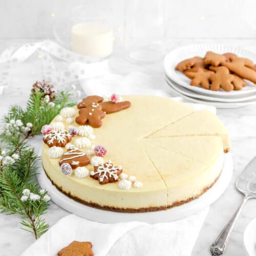 pulled back shot of eggnog cheesecake on white plate with three slices cut, pine branches beside, a plate of cookies, glass of eggnog, and white ribbon behind