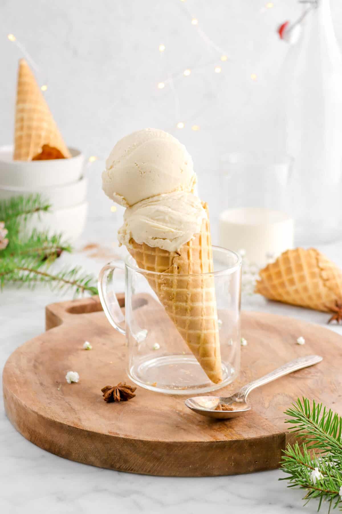 tilted cone with eggnog ice cream on wood serving board with spoon, star anise pod, pine branches, bowls, and mug of eggnog behind