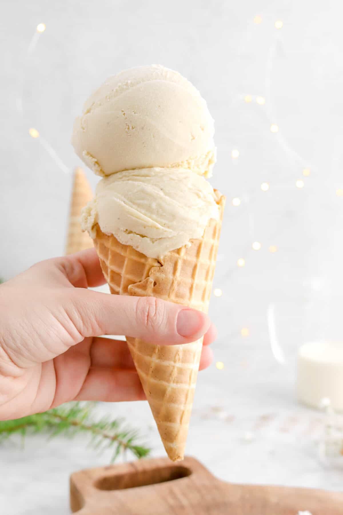 hand holding ice cream cone with two scoops of eggnog ice cream with fairy lights behind