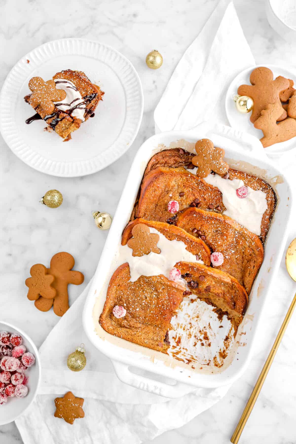 overhead shot of gingerbread french toast casserole on white napkin with slice of casserole on white plate beside, cookies and mini gold ornaments around