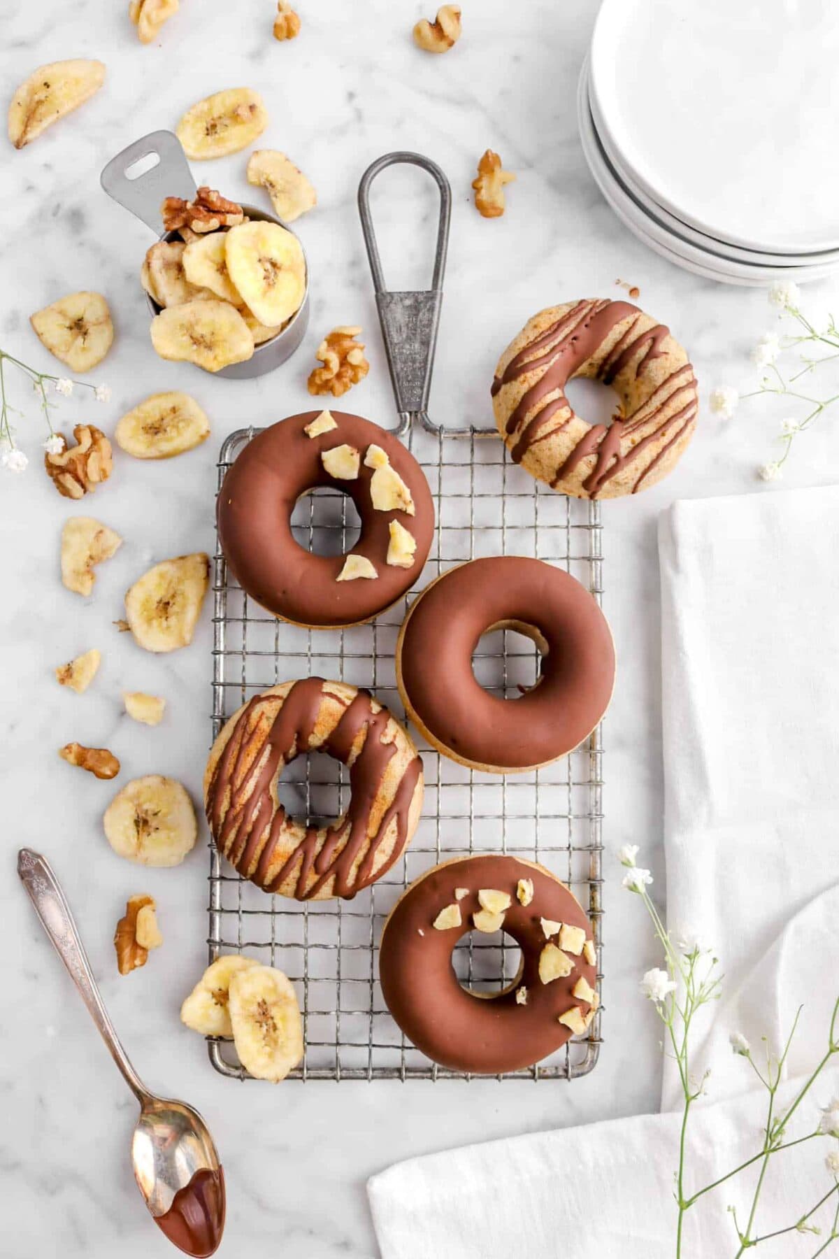 overhead shot of five donuts with banana donuts with banana chips, walnuts, stack of white paltes, and flowers