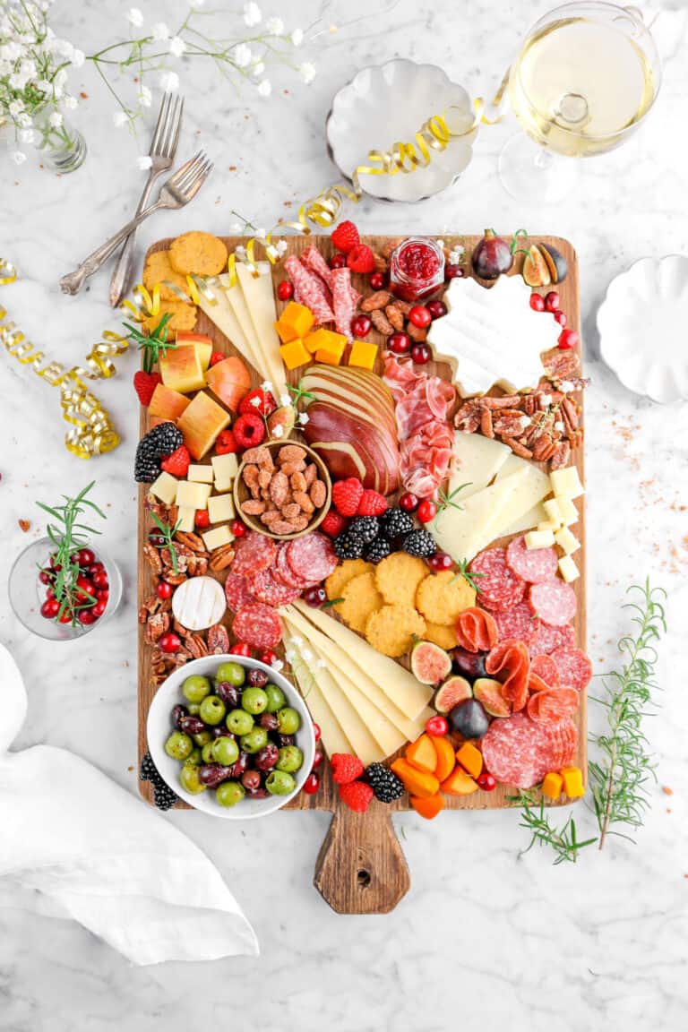 overhead shot of charcuterie board with glass of champagne, forks, rosemary sprigs, flowers, and white napkin