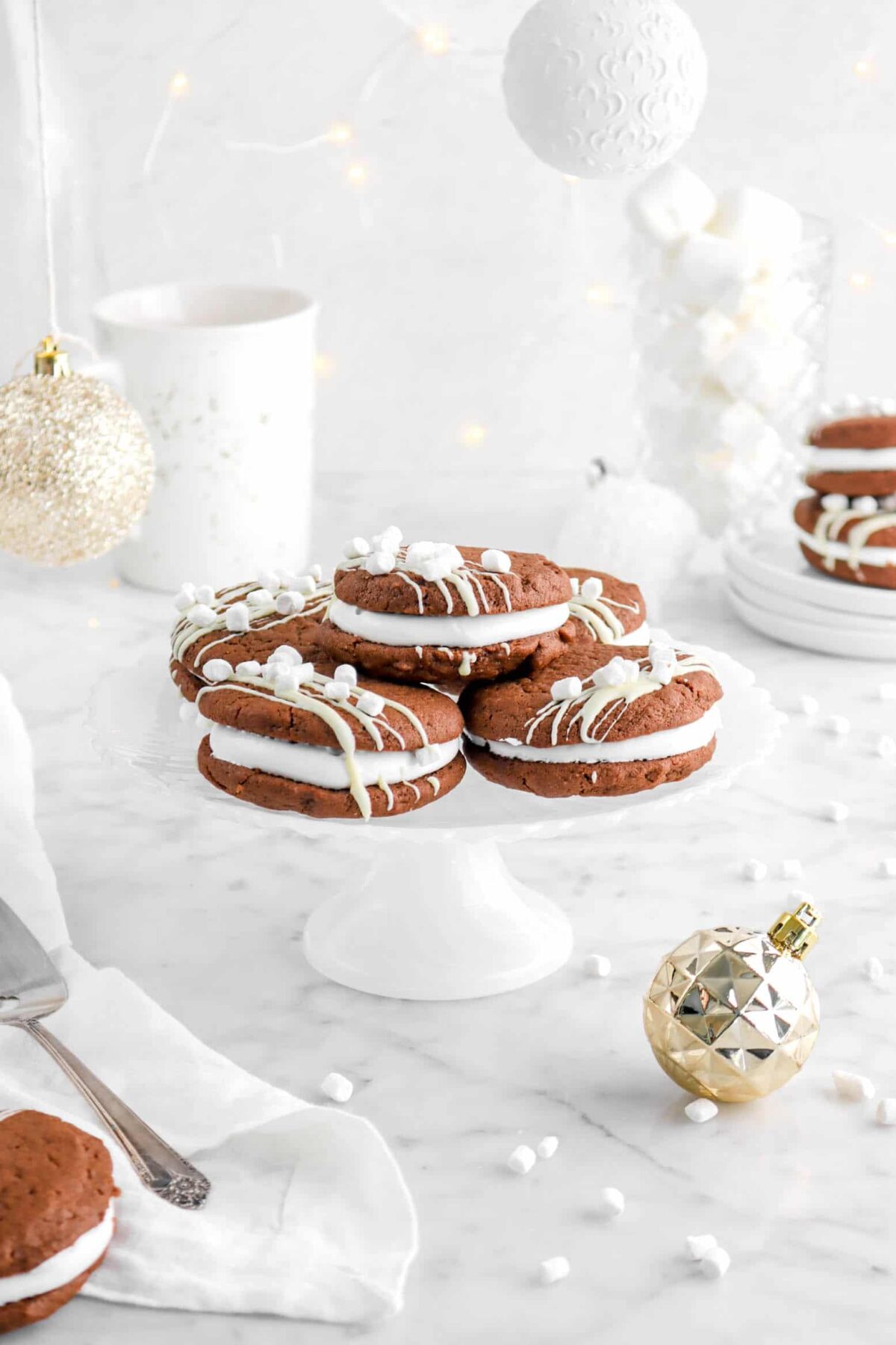 hot cocoa sandwich cookies on white milk glass cake plate with white and gold ornaments, a white napkin, a mug, fairy lights, and mini marshmallows