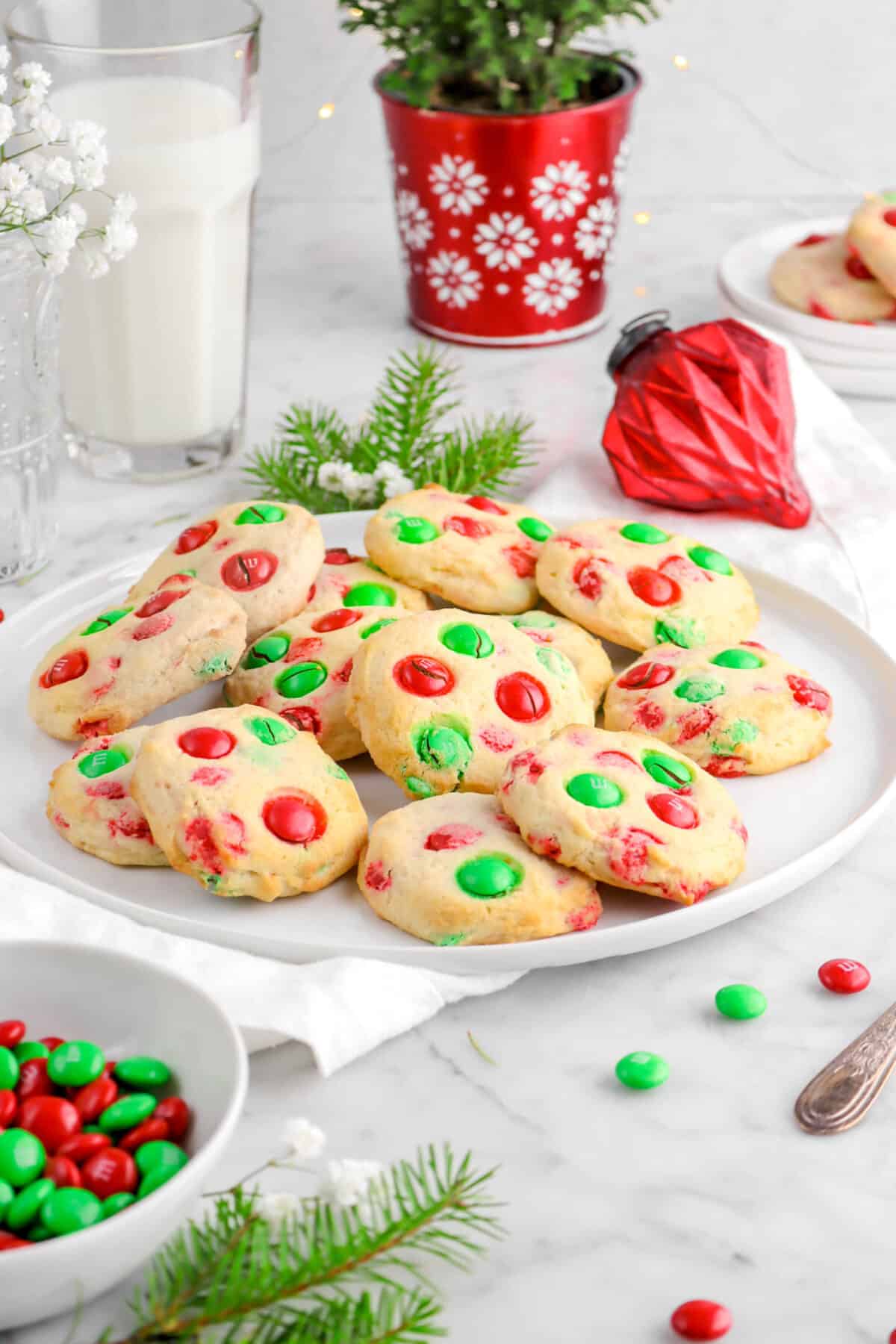 m&m cookies on white plate with red ornament behind, glass of milk, and ever green branches