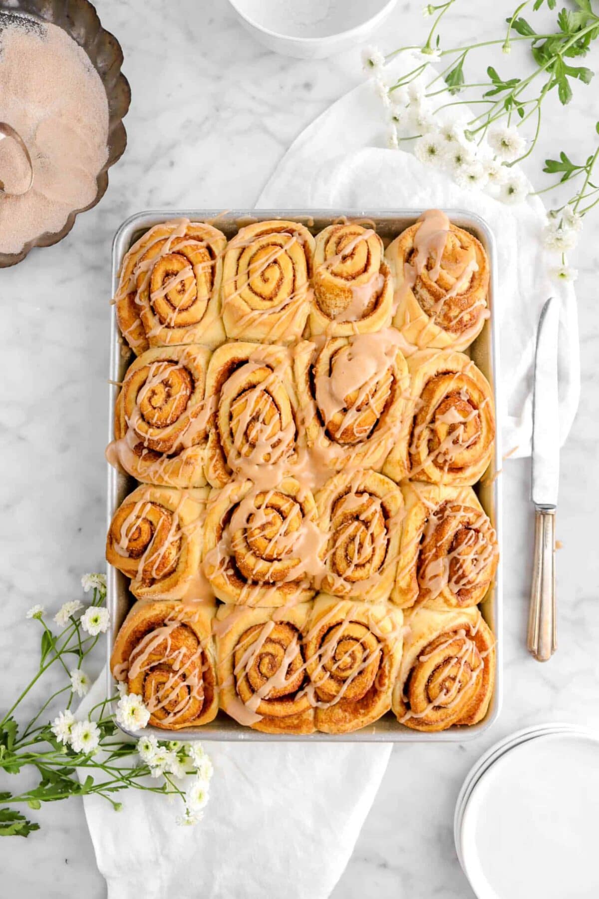 overhead shot of cinnamon rolls in sheet pan with icing, white chamomile flowers, a knife, and plates