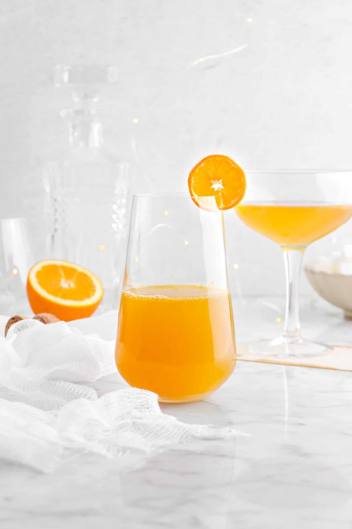 orange champagne cocktail with glass behind, half of an orange, and white cheese cloth