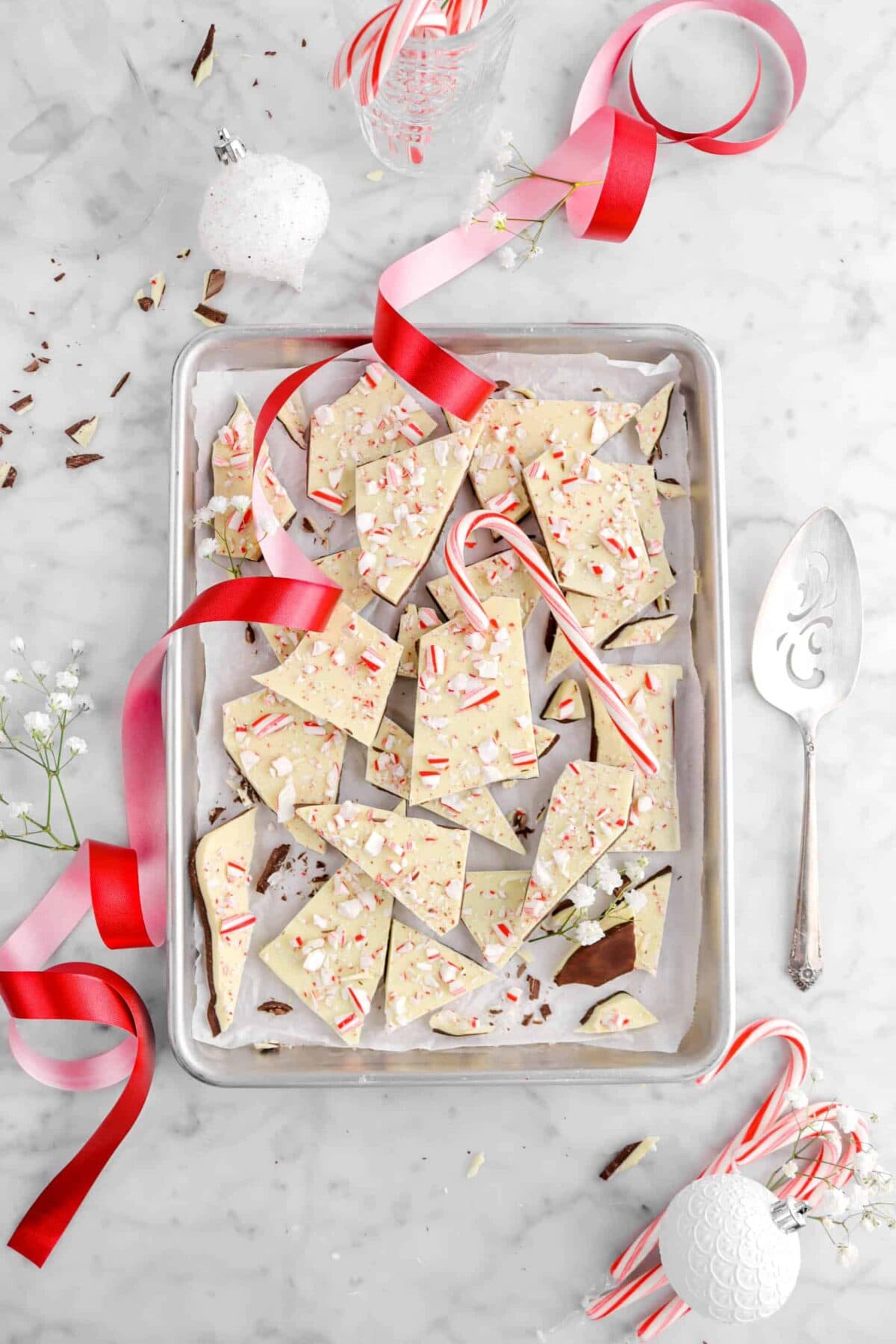 peppermint bark on sheet pan with red ribbon, white ornaments, white flowers, and candy canes