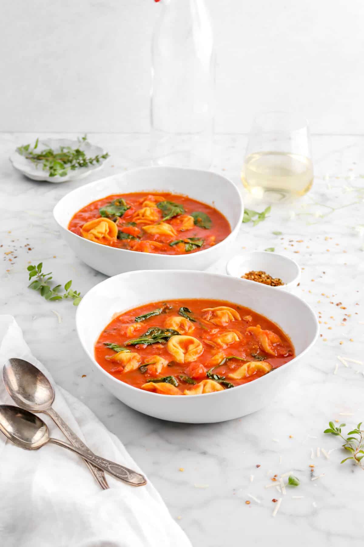 two bowls of tomato tortellini soup on counter with two spoons with fresh herbs, glass of wine, and red pepper flakes around