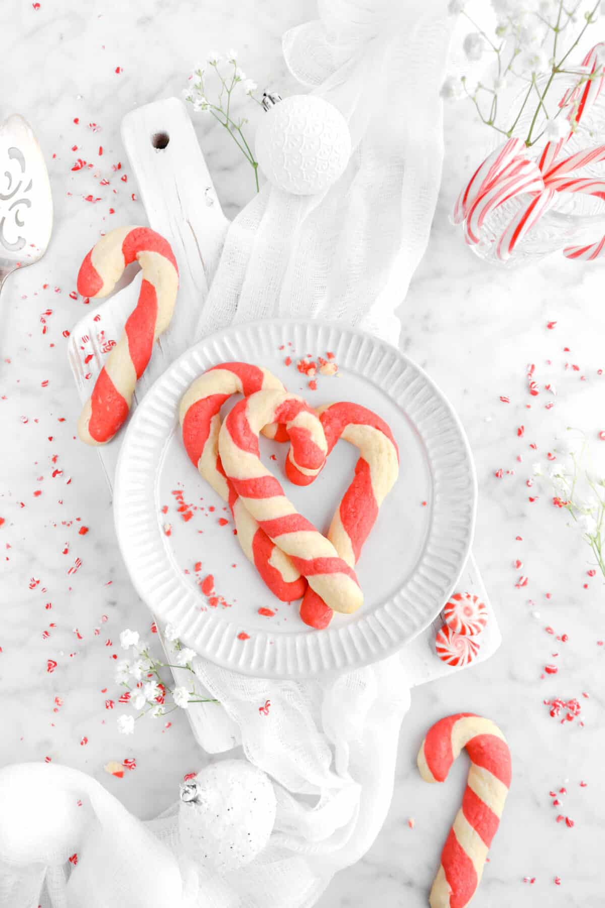 three candy cane cookies on white palte on serving board with candy canes, peppermint candies around, with flowers