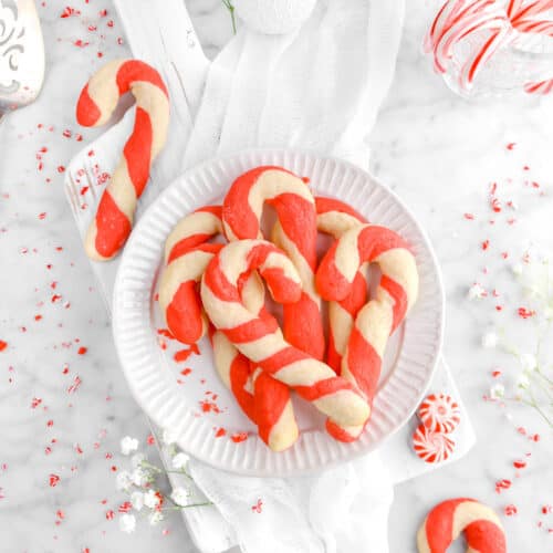 plate full of candy cane cookies on white wood board with two other candy cane cookies, peppermint candies, and flowers