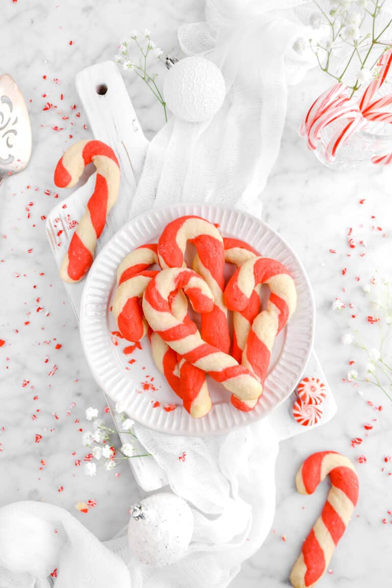 plate full of candy cane cookies on white wood board with two other candy cane cookies, peppermint candies, and flowers