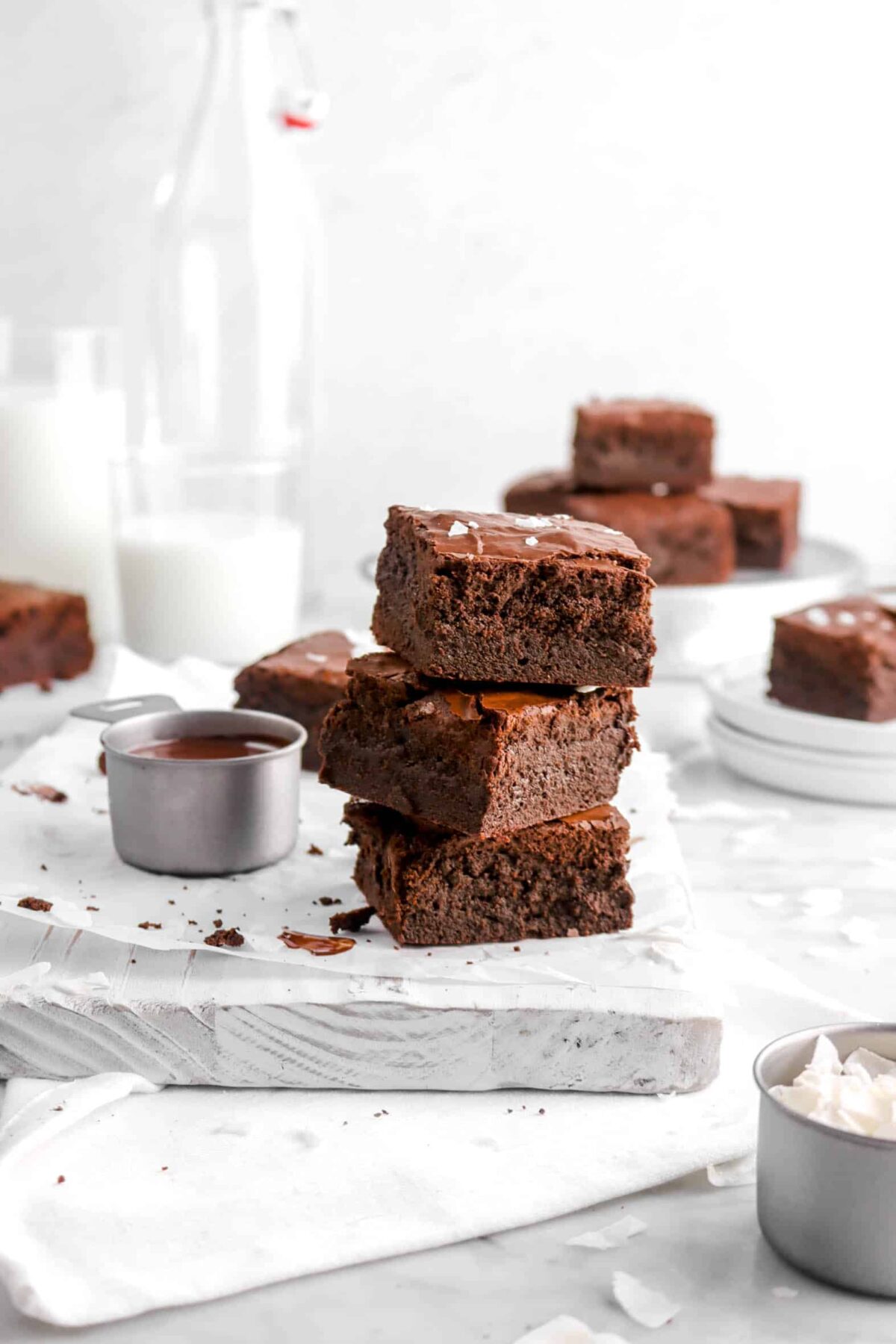 three stacked brownies on board with melted chocolate, two glasses of milk behind, and coconut flakes around