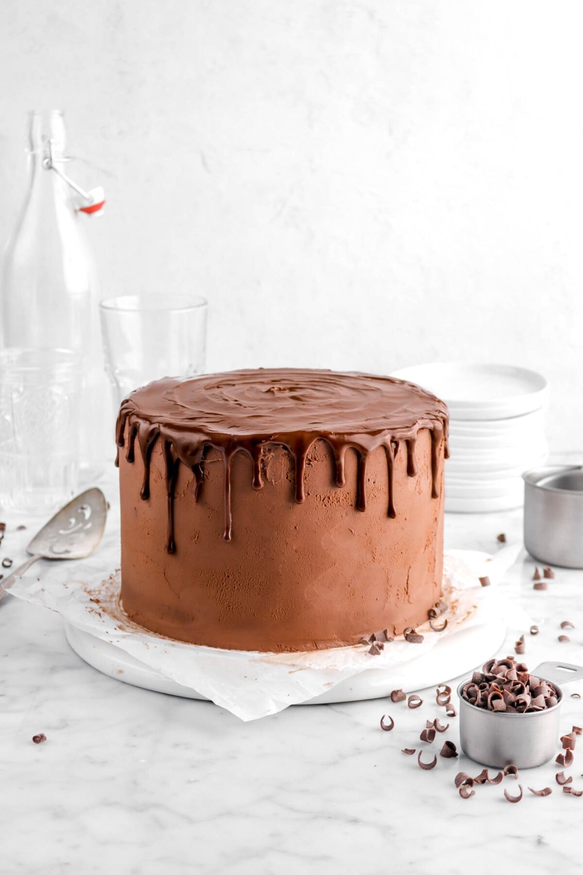 chocolate cake on upside down white plate with parchment paper, chocolate curls and empty glasses behind