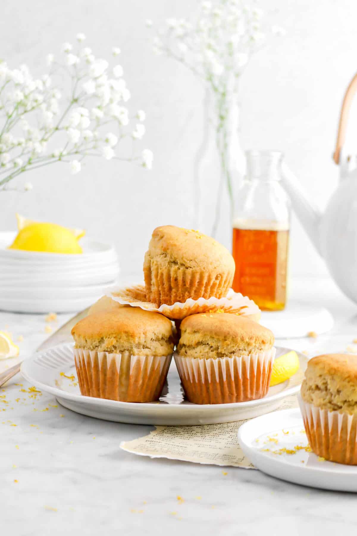 three stacked muffins white plate with glass of honey, tea pot, flowers, and stack of white plates behind