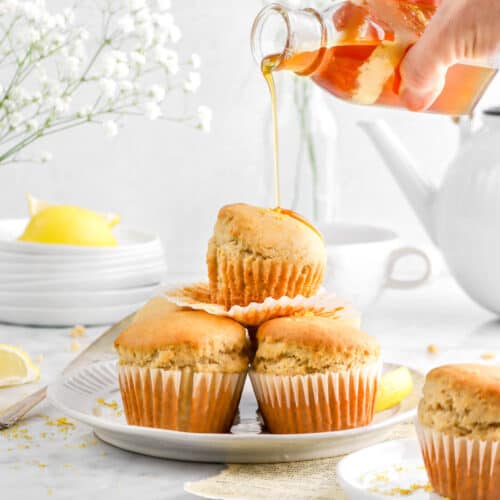 honey being poured onto stack of muffins on white plate with another muffin in front, a stack of paltes behind, flowers, and white teapot