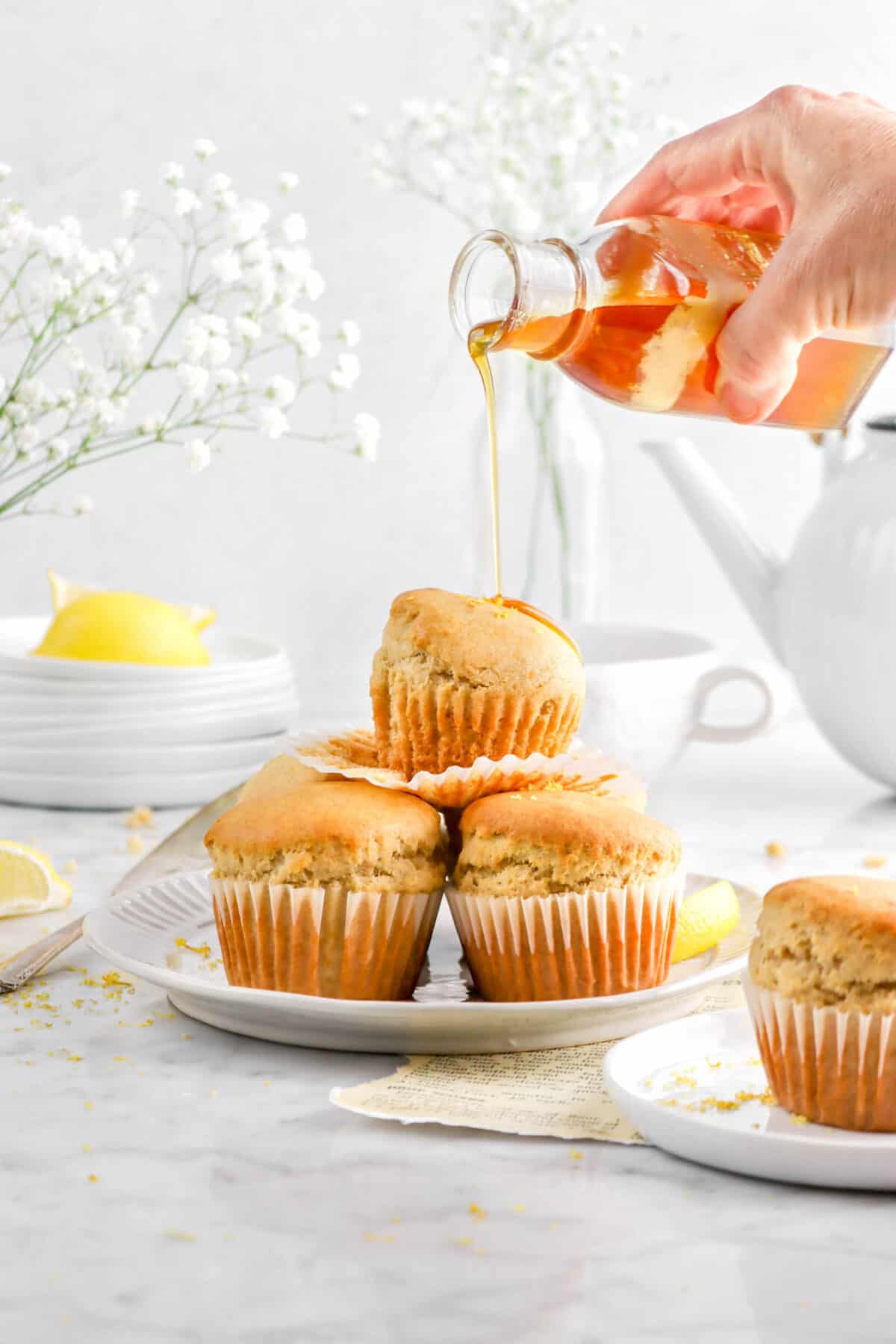 honey being poured onto stack of muffins on white plate with another muffin in front, a stack of paltes behind, flowers, and white teapot