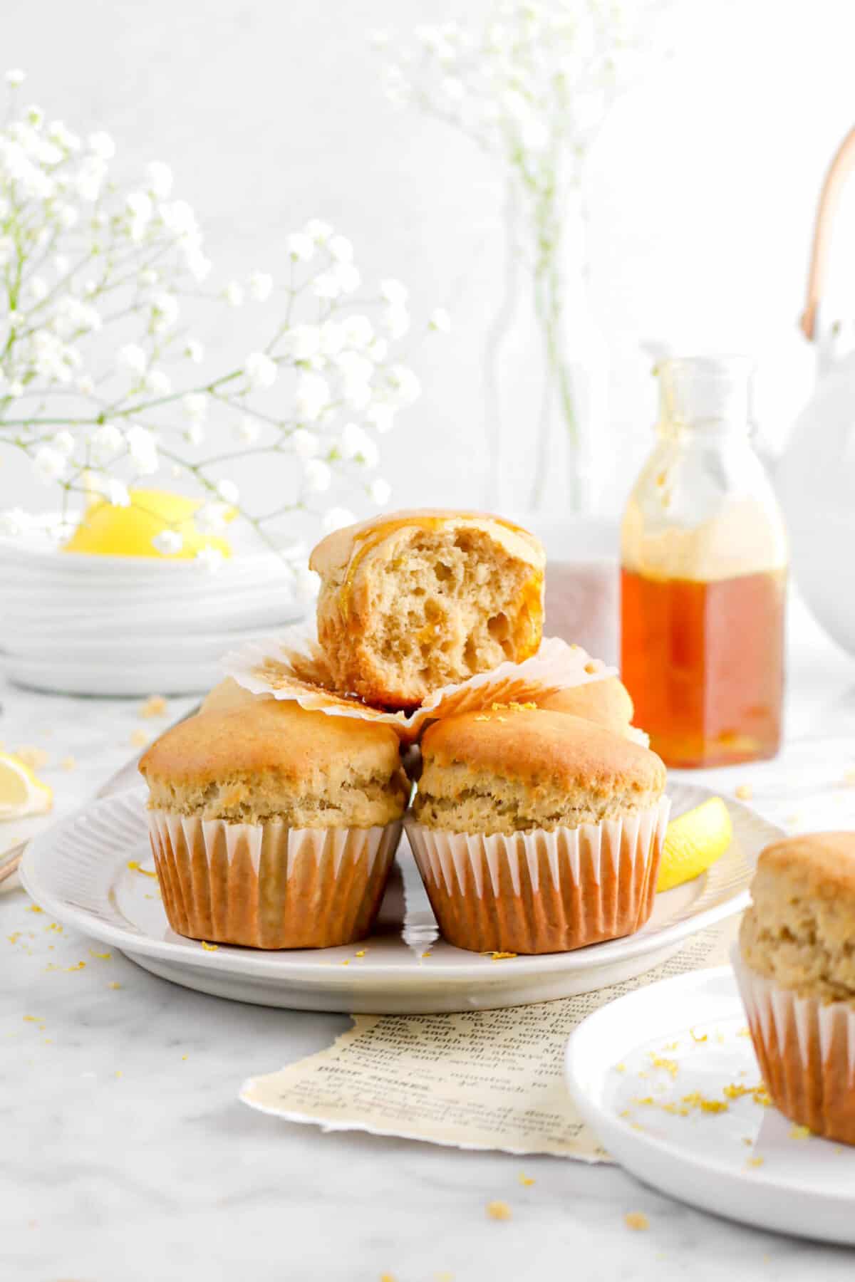 earl grey honey lemon muffins stacked white plate with glass of honey behind, a tea pot, a stack of white plates, and flowers in vases