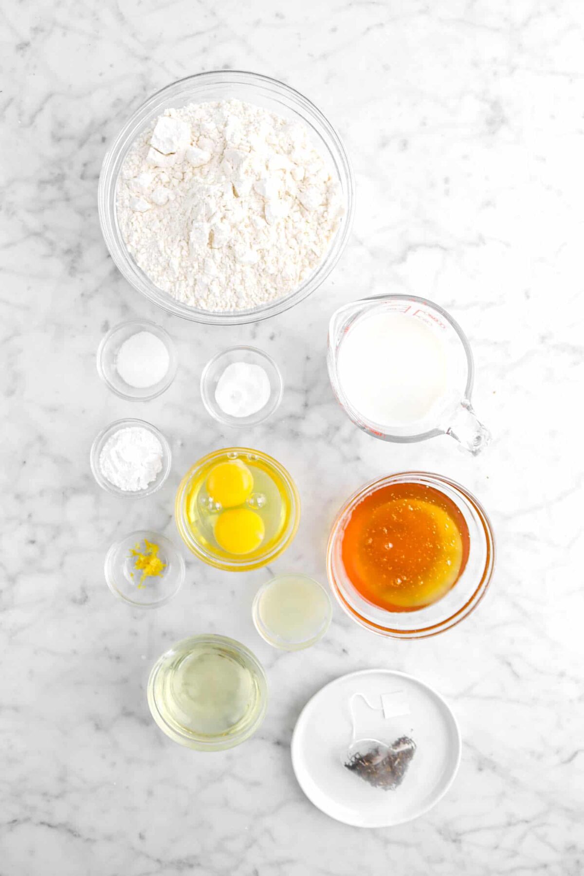 flour, salt, baking soda, baking powder, eggs, lemon zest, lemon juice, vegetable oil, tea bag, honey, and milk on marble counter