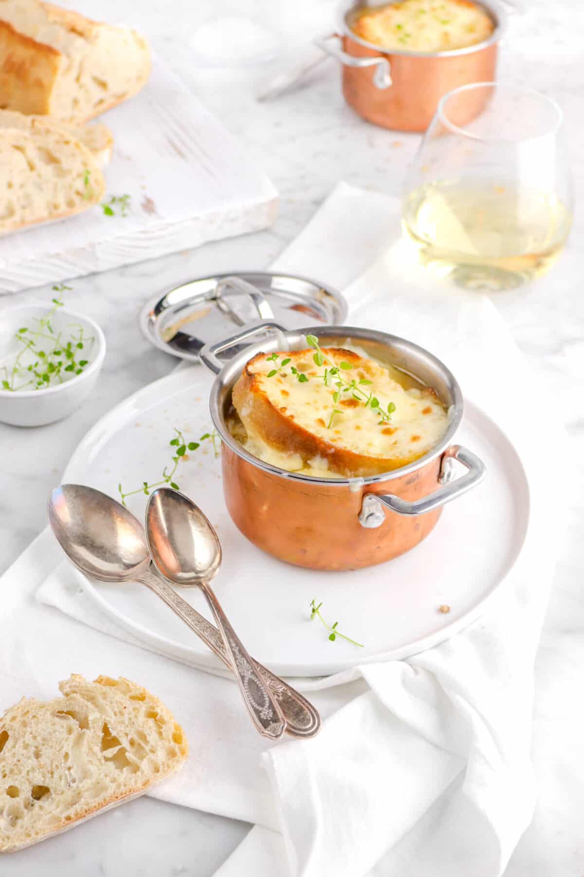 french onion soup in copper plate on white plate with another pot behind and glass of white wine
