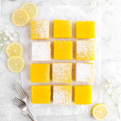overhead shot of twelve lemon bars on parchment paper with flowers, lemon slices, forks, stack of white plates, and measuring cup full of powdered sugar
