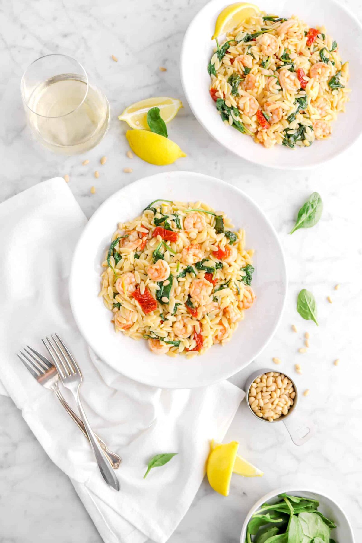 two bowls of shrimp orzo on marble counter with white napkin, two forks, spinach leaves, pine nuts, lemon wedges, and glass of wine