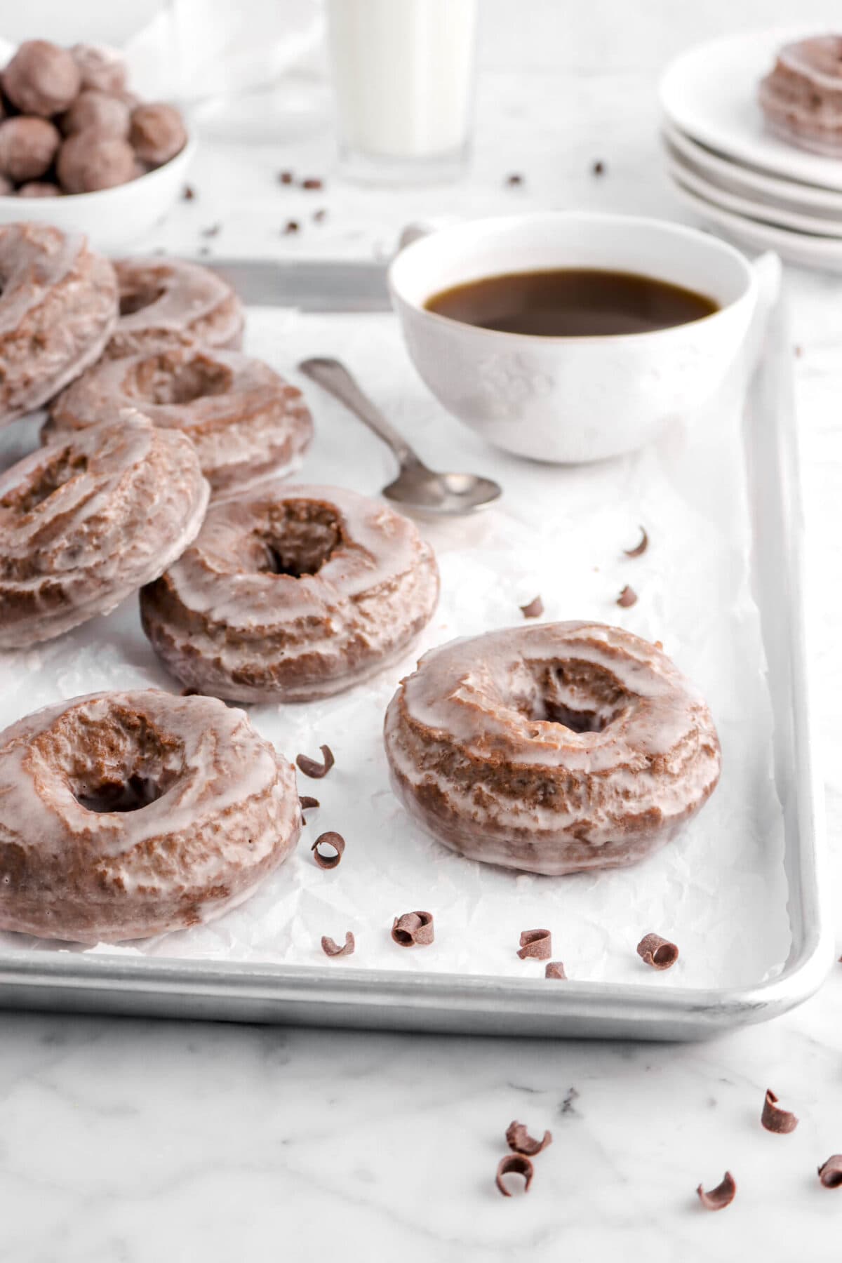 chocolate doughnut on parchment paper in sheet pan with more doughnuts behind and mug of coffee