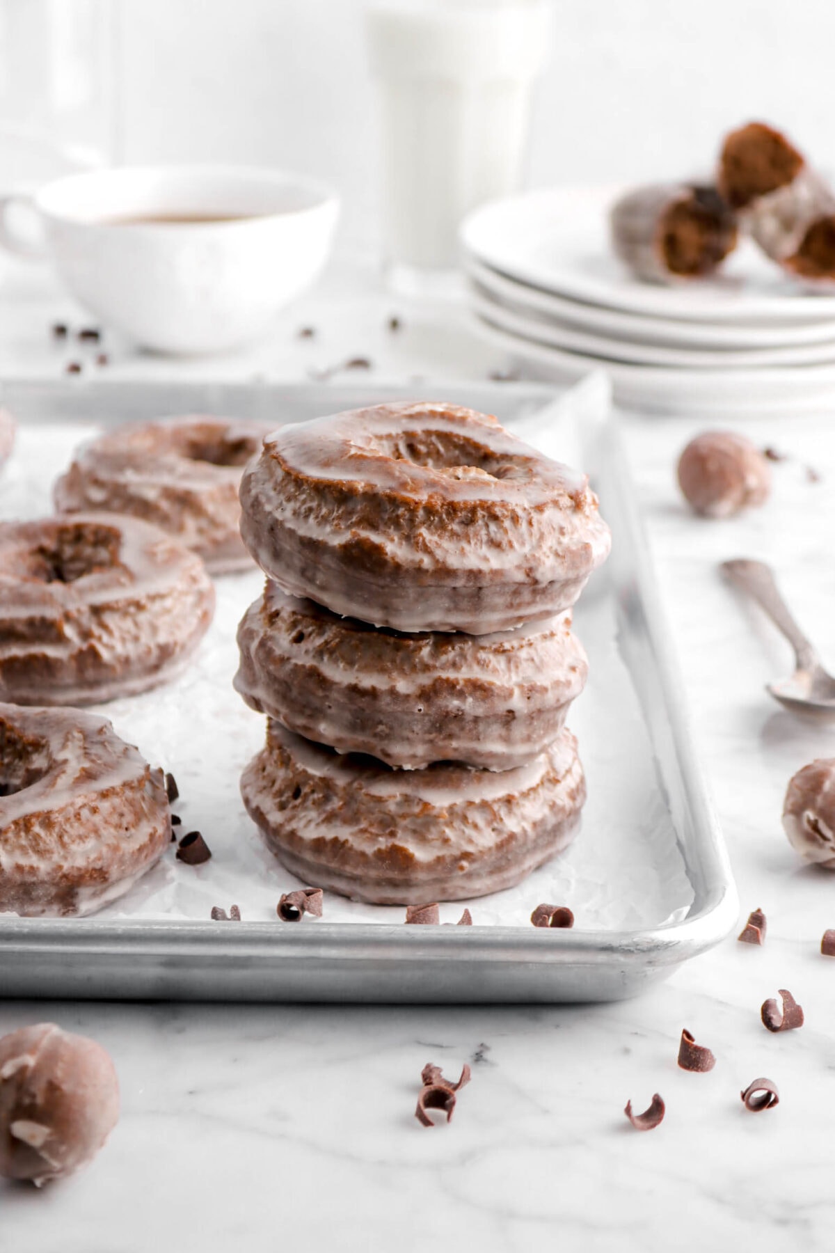 three stacked chocolate doughnuts on sheet pan with mug of coffee, glass of milk, and a broken doughnut behind on stacked plates