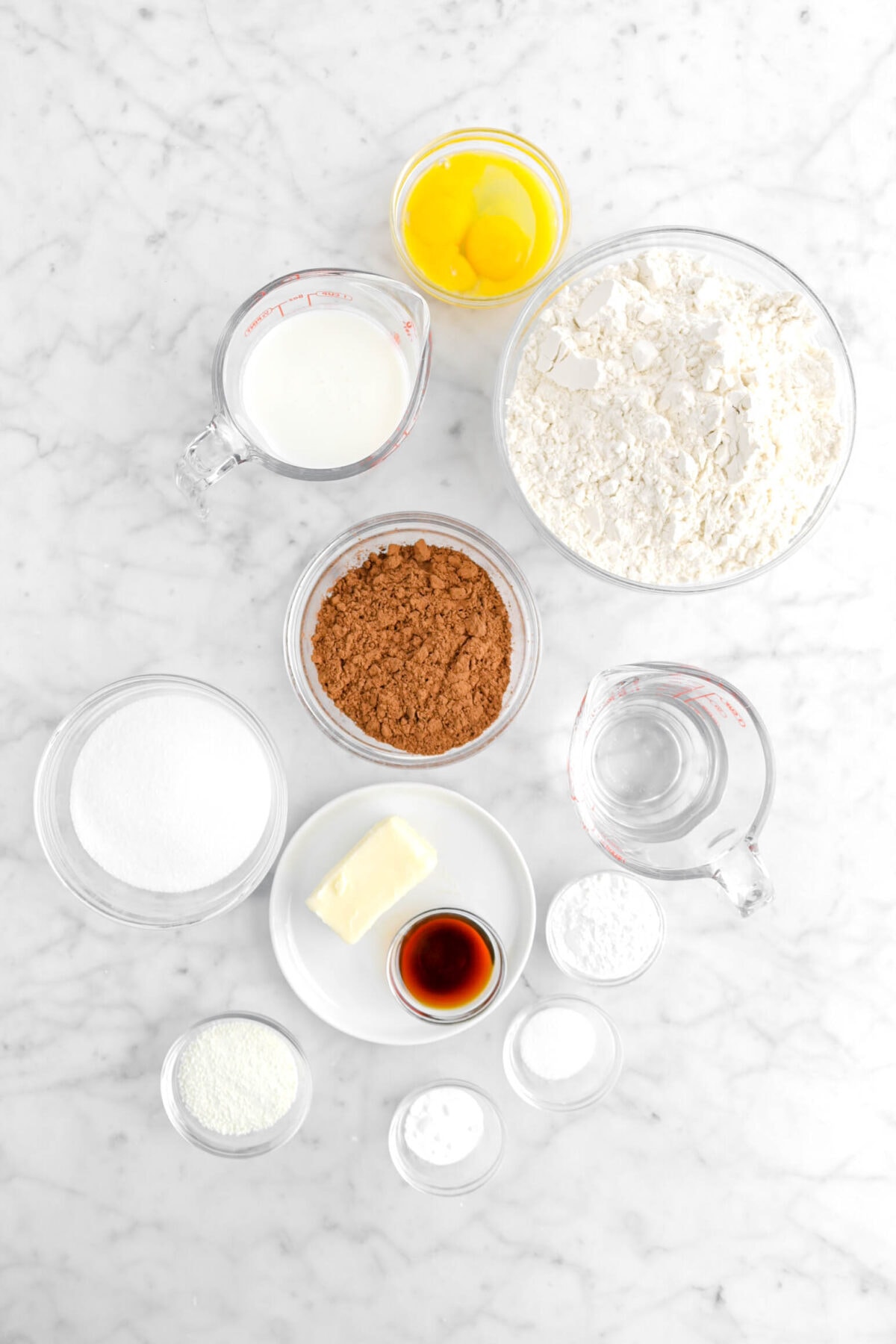 eggs, milk, flour, cocoa powder, water, butter, vanilla, sugar, milk powder, baking soda, salt, and baking powder on marble counter