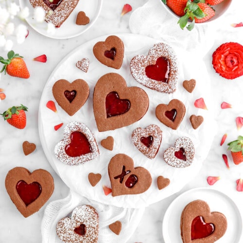 flat lay of chocolate linzer cookies with strawberry filling, fresh strawberries, red flowers, and white cheese cloth