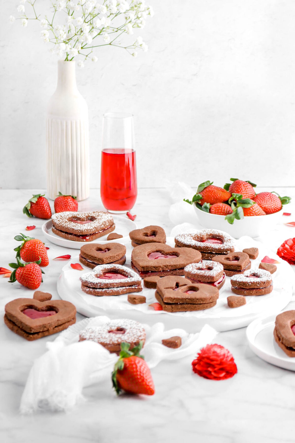 front shot of chocolate linzer cookies with strawberries, glass of wine, vase of flowers, and white cheese chloth