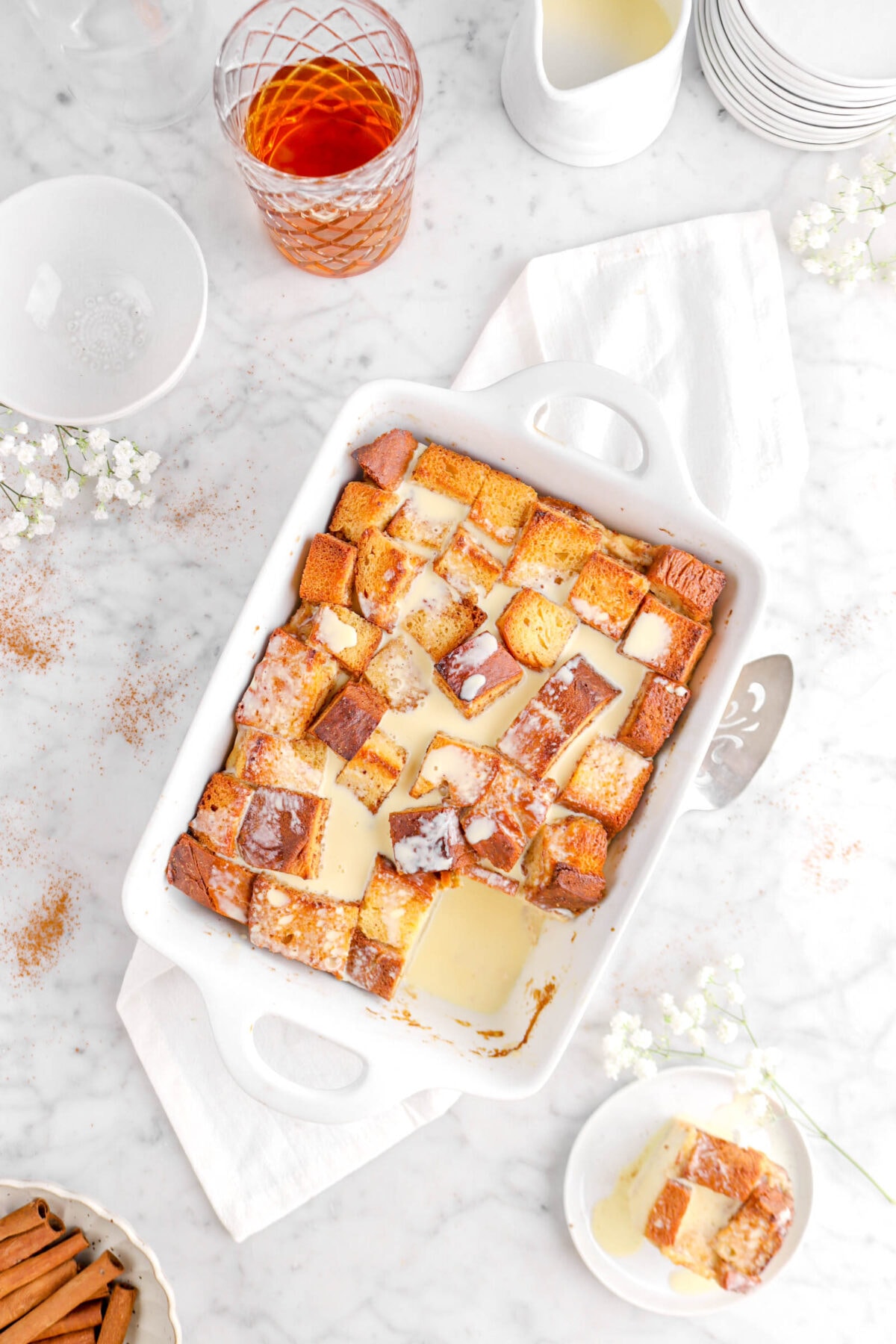 pulled back flat lay of bourbon bread pudding on white napkin with slice on white plate with flowers, cake knife, glass of bourbon, and stack of plates