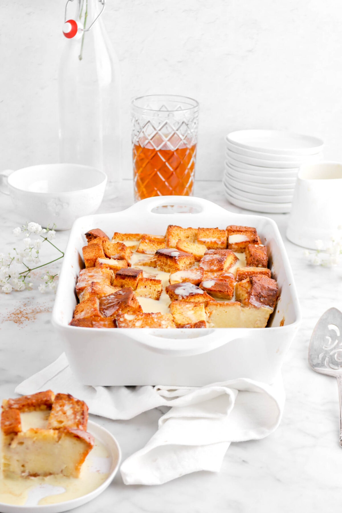 front shot of bread pudding casserole with slice on white plate in front, a cake knife, flowers, glass of bourbon, stack of plates, and coffee mug behind