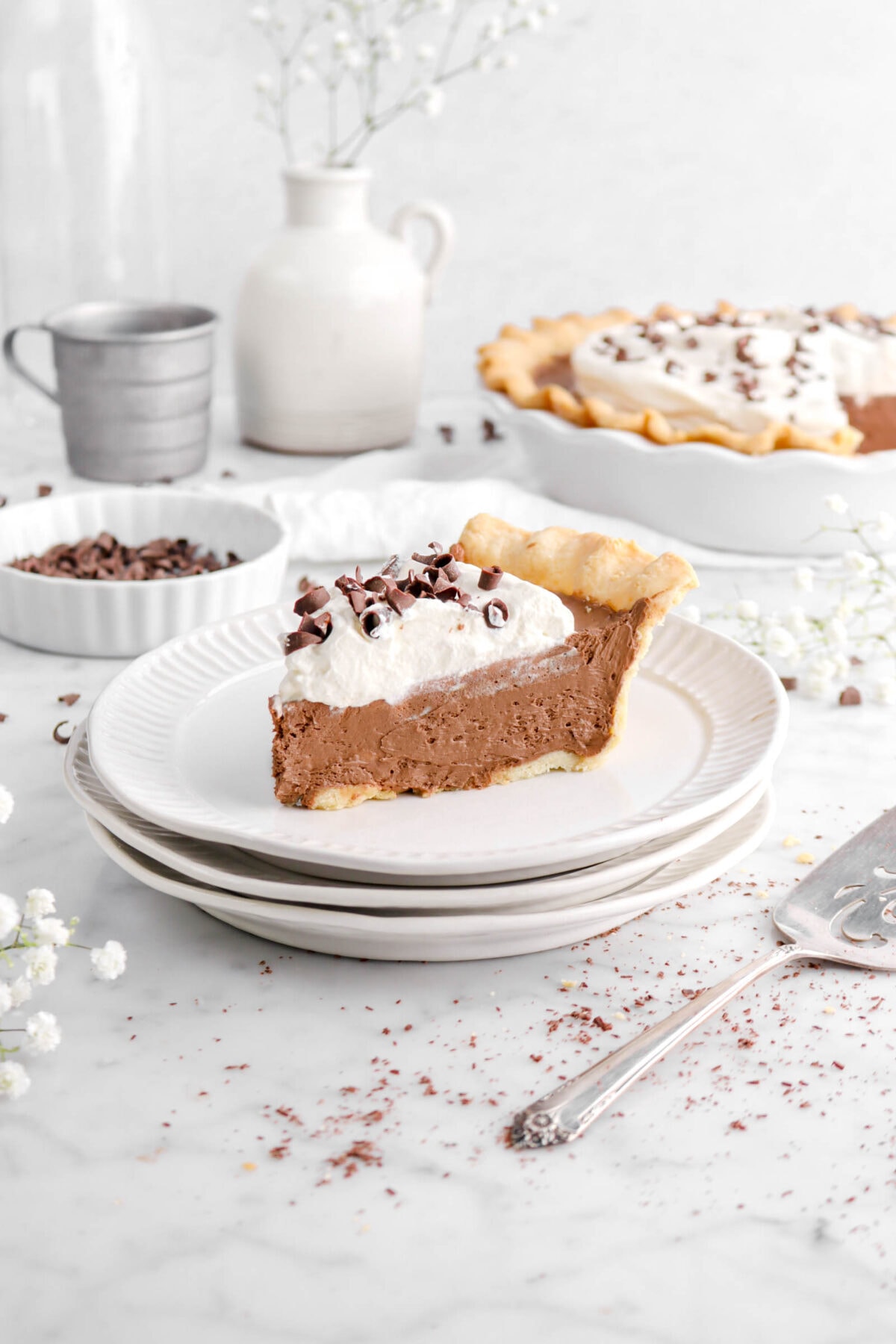 pulled back shot of slice of french silk pie on marble counter with bowl of chocolate curls, metal measuring cup, white vase, and whole pie behind