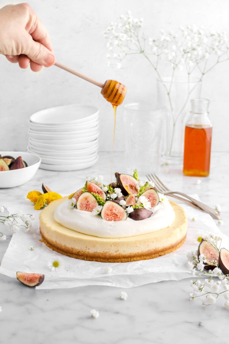 honey being drizzled over sheet cheesecake with two forks, jar of honey, fresh figs, and flowers around