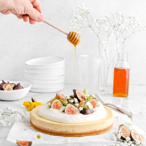 pulled back shot of honey being drizzled over sheet cheesecake with two forks, jar of honey, fresh figs, and flowers around