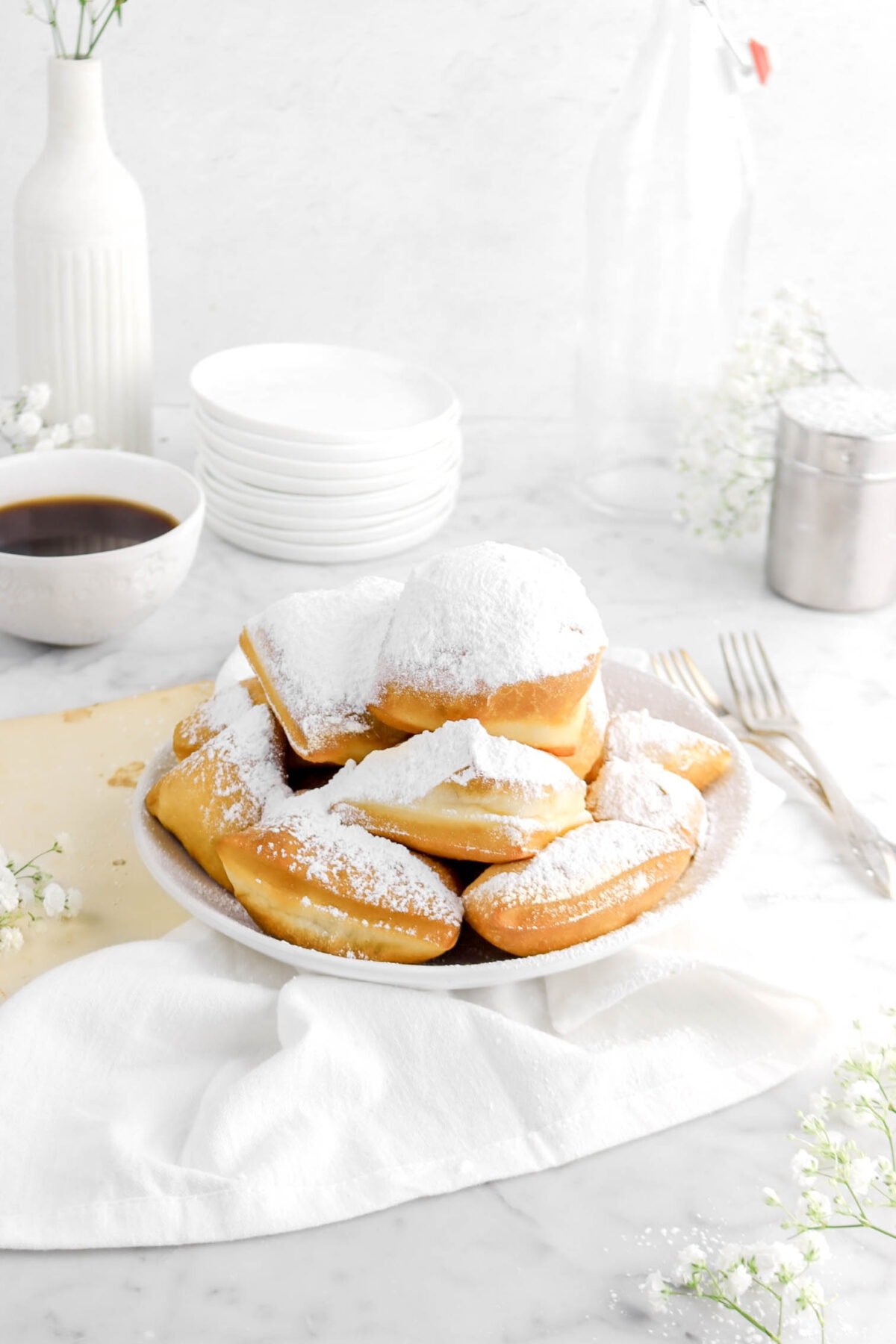 angled photo of beignets in white bowl withs tack f plates, flowers, cup of coffee, forks, and white napkin