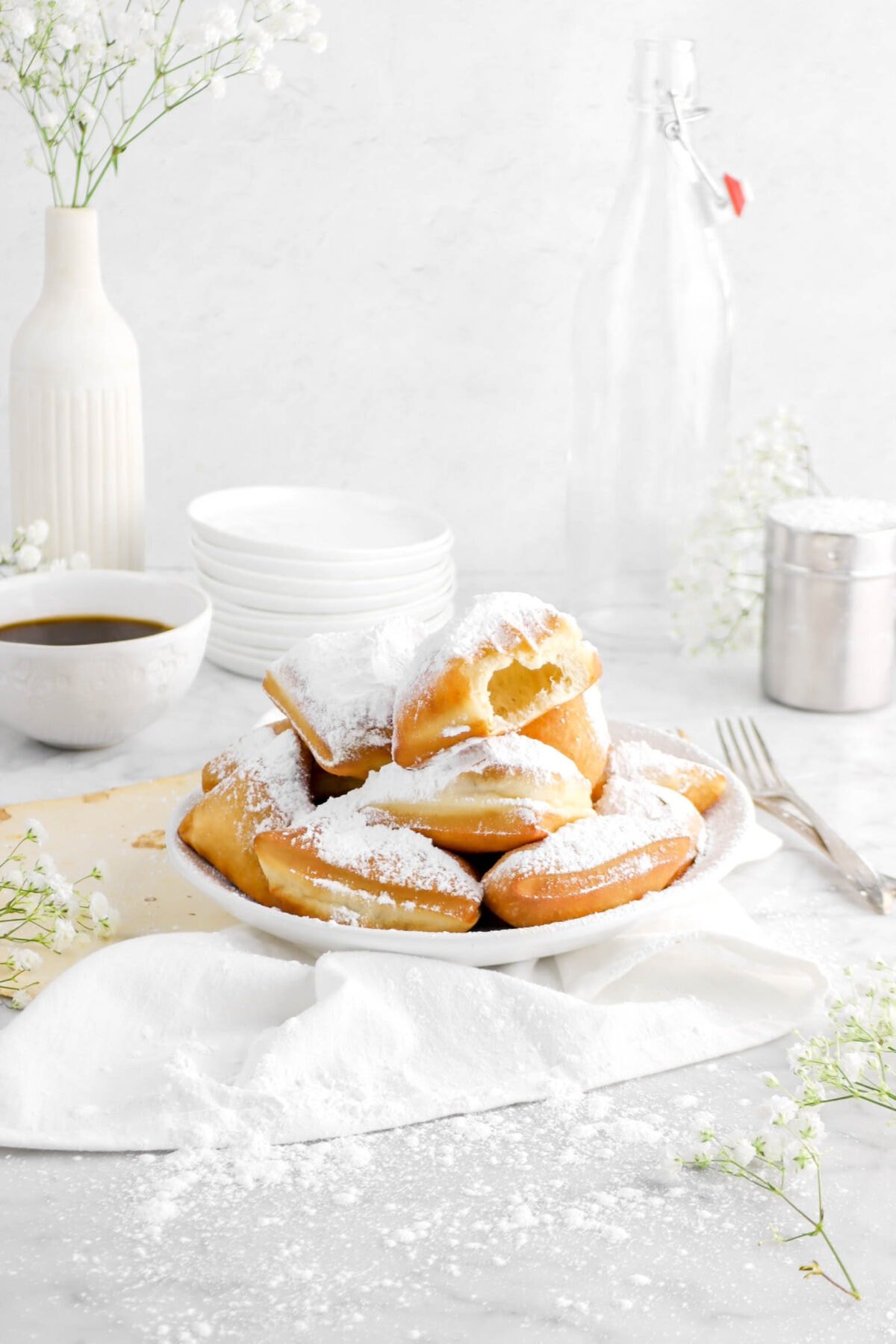 angled photo of beignets in white bowl with bite missing from top beignet on white napkin with old book pages, two forks, and stack of white plates behind