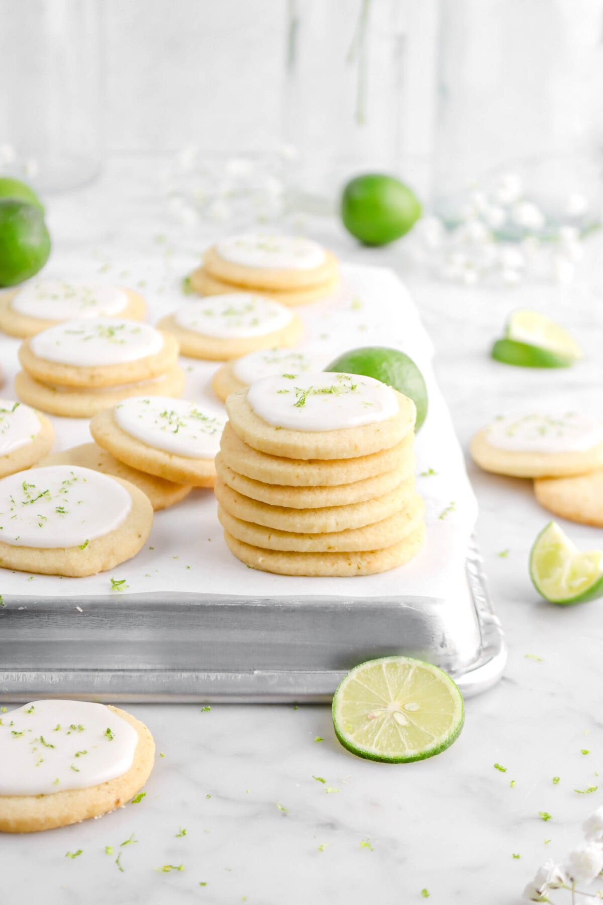 stacked key lime cookies on upside down sheet pan with more cookies behind, key limes, and white flowers on marble surface