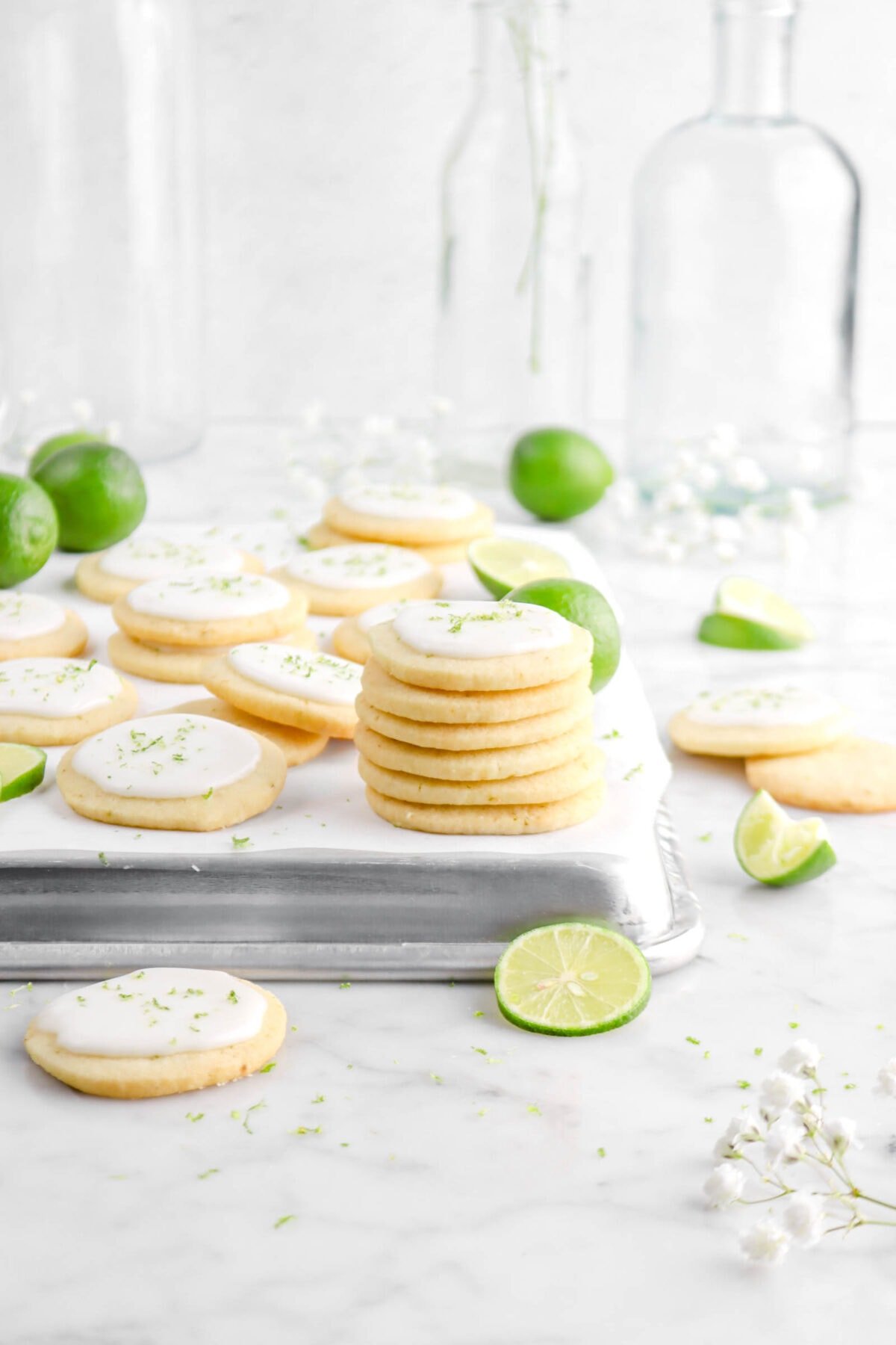 front shot of stacked key lime shortbread cookies on upside down pan with more cookies around, key limes, white flowers, and empty glass jars behind