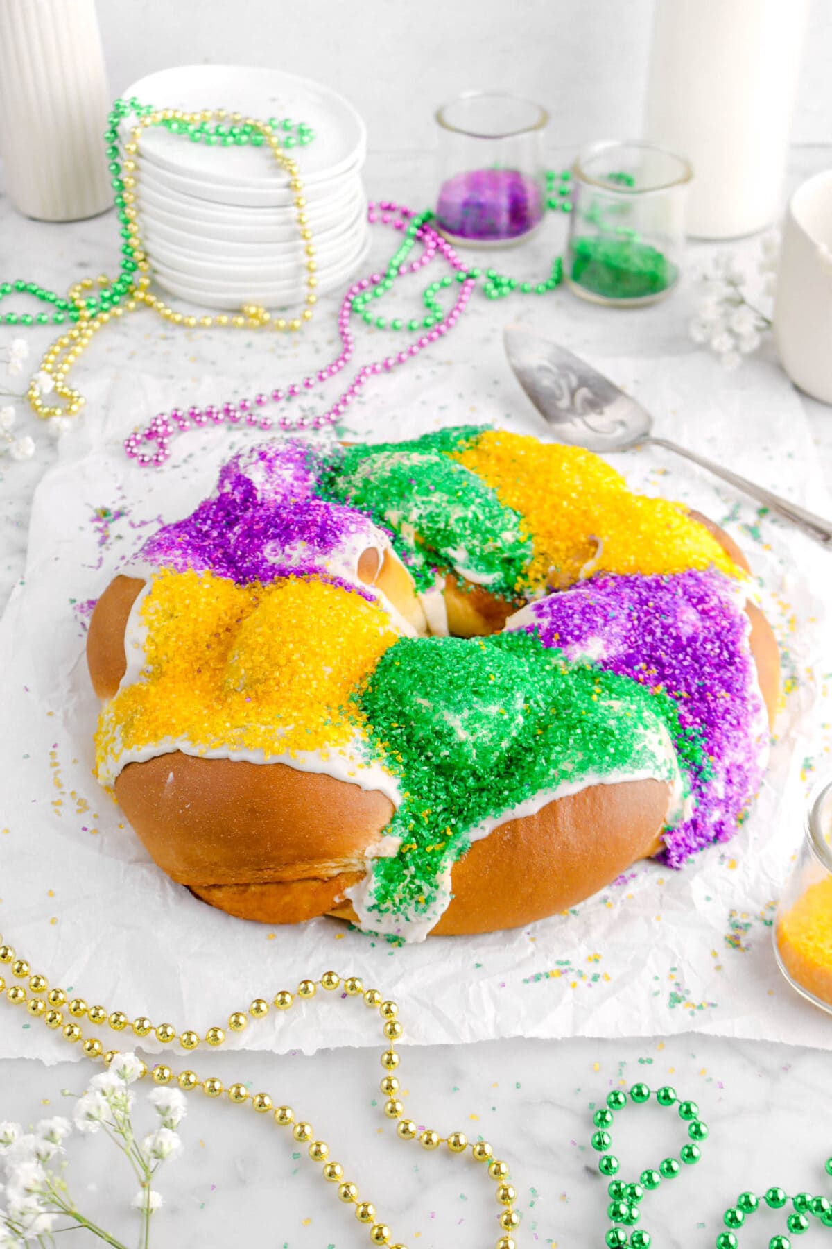 angled close up of king cake on parchment paper with mardi gras beads and decorative sugar around