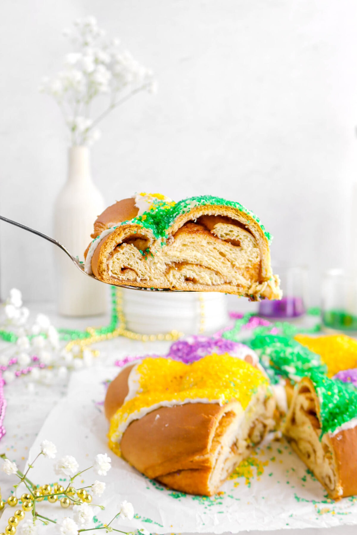 slice being held over king cake on cake server with flowers and plates behind