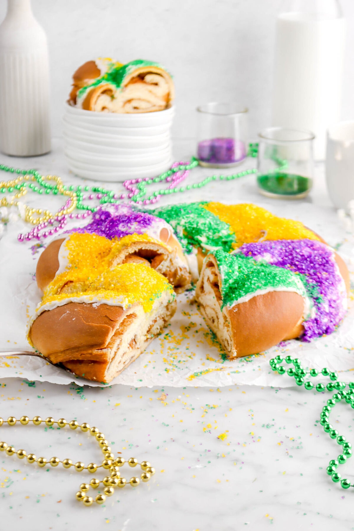 front shot of king cake on parchment paper with slice on cake server, slice on stack of plates behind, and mardi gras beads around