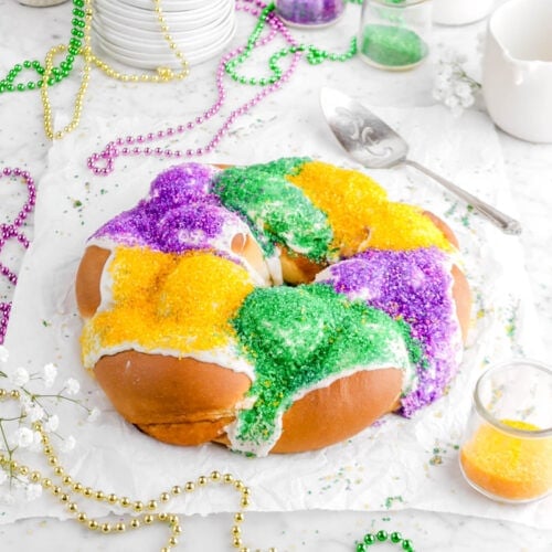 angled shot of king cake on parchment paper with flowers, beads, decorative sugar in jars, and glass of milk