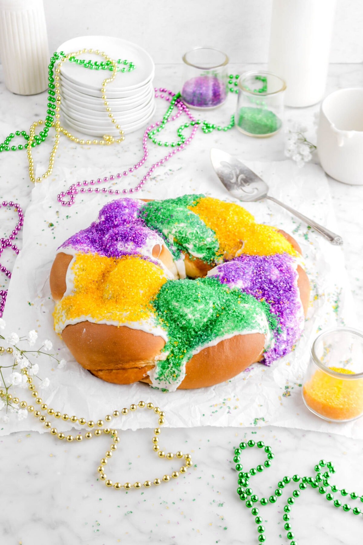 angled shot of king cake on parchment paper with flowers, beads, decorative sugar in jars, and glass of milk