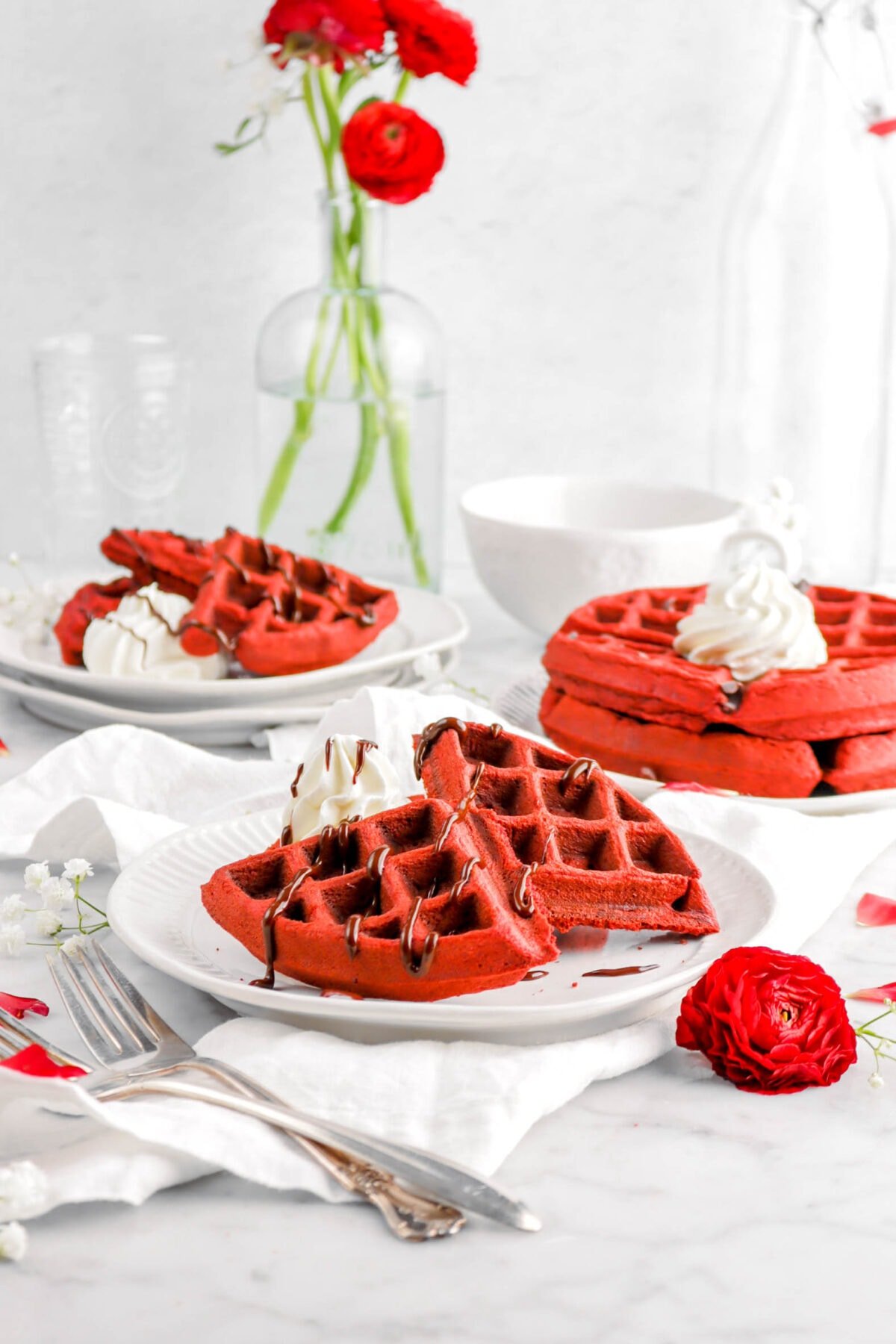 three plates of red velvet waffles on white plates with red flowers, two forks, and a white napkin