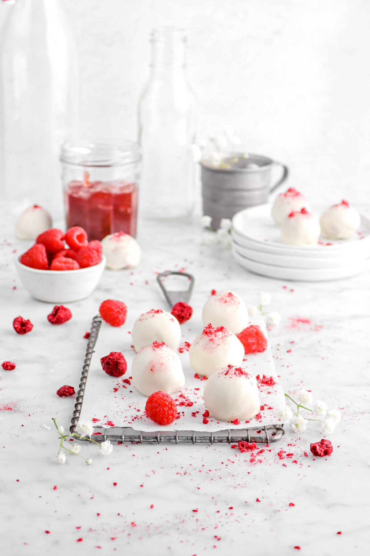 five white chocolate raspberry truffles on parchment paper with jar of jam, bowl of fresh raspberries, stacked plates, and empty glasses behind